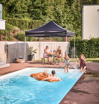 The black folding gazebo stands by the pool in the garden. Friends sit under it in the shade and have a pool and garden party.