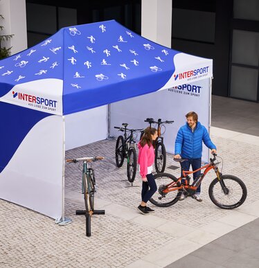 The promotion tent from Intersport is a 4.5x3 m folding gazebo. It is printed all over in white and blue. Various sport icons can be seen on the roof. The salesman shows the customer the bicycle.