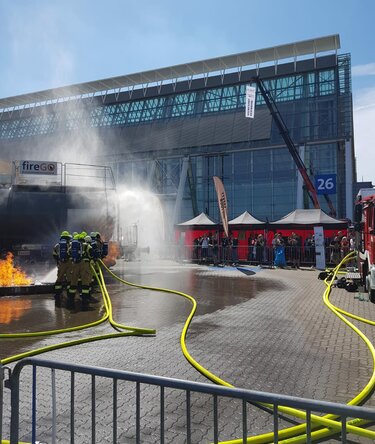 Firefighters during an outdoor firefighting demonstration at a trade fair, with a water jet, emergency vehicles, and emergency tents in the background.